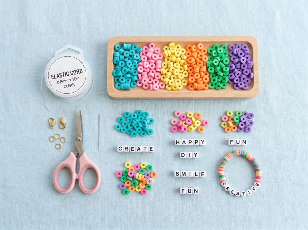 A flat lay of beaded jewelry making supplies for beginners, featuring clear elastic cord, colorful clay beads, and letter beads.
